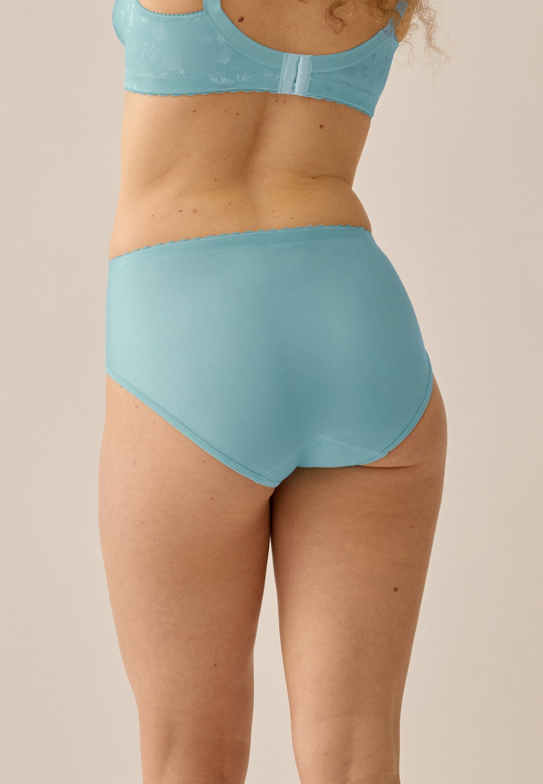 Achterzijde van vrouw die de minimizer slip met bloemenpatroon in de kleur aquamarine  van het merk Naturana draagt